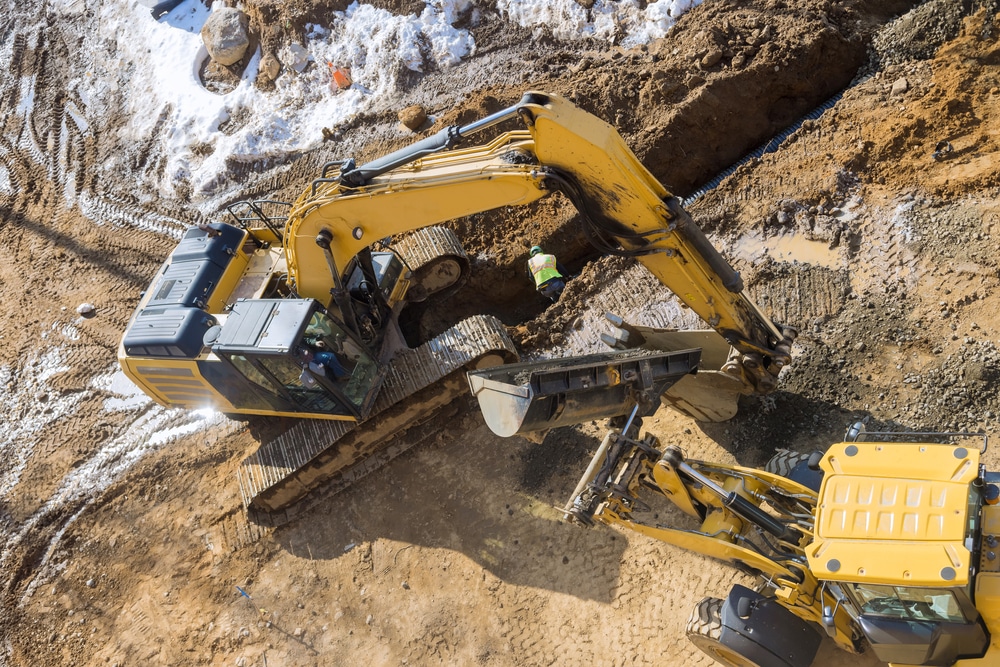 Excavatrice sur un chantier de construction : terrassement Excavatrice jaune creusant une tranchée sur un chantier de construction boueux avec un ouvrier et une chargeuse.