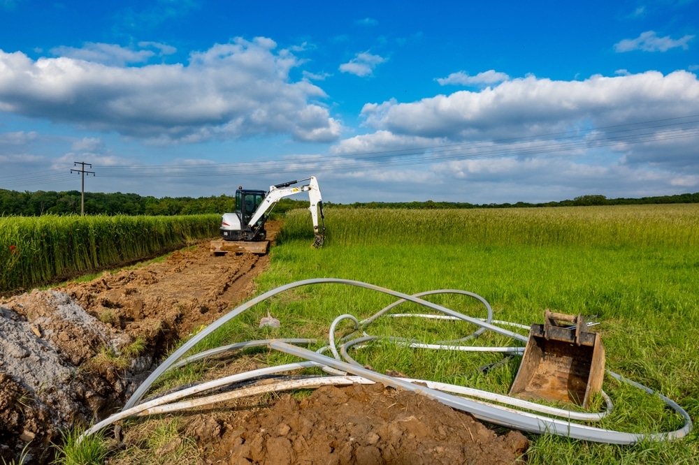 Installation de tuyaux par mini-pelleteuse dans un champ Mini-pelleteuse creusant une tranchée pour des tuyaux dans un champ agricole sous un ciel bleu nuageux.