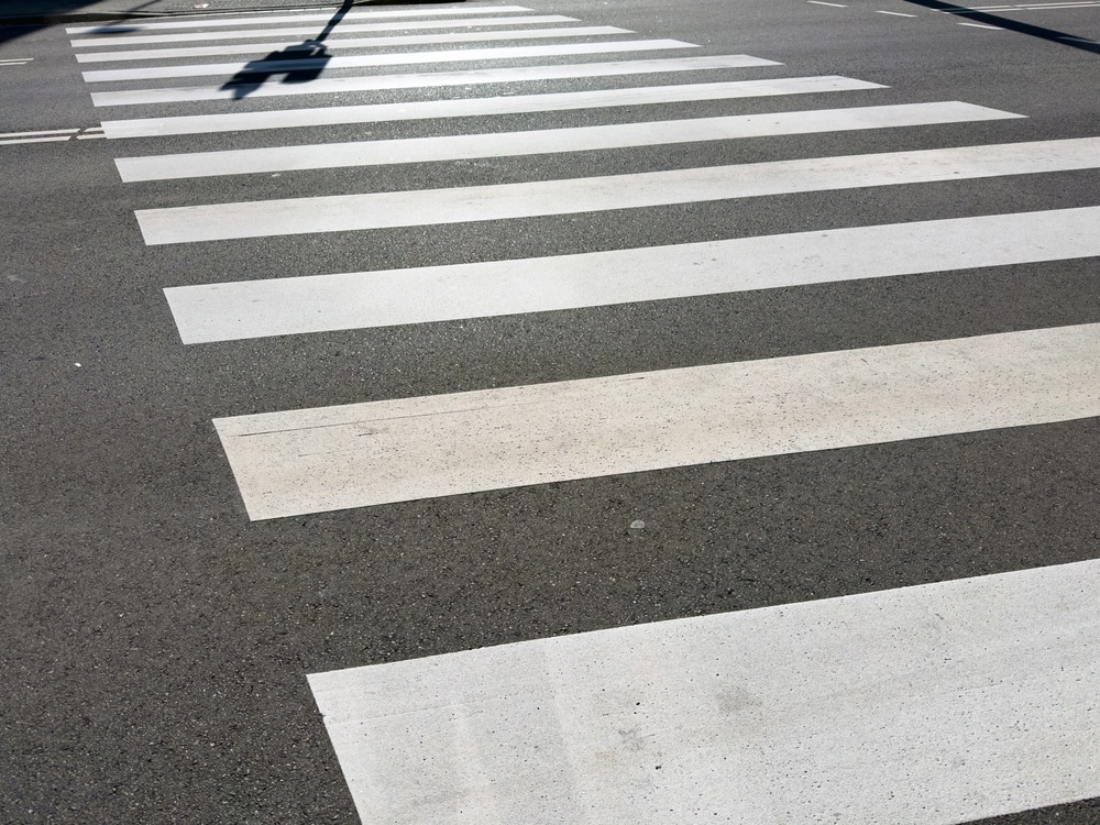 Passage Piéton : Sécurité Routière et Marquage au Sol Passage piéton aux larges bandes blanches sur bitume sombre avec ombre portée d'un poteau de signalisation.