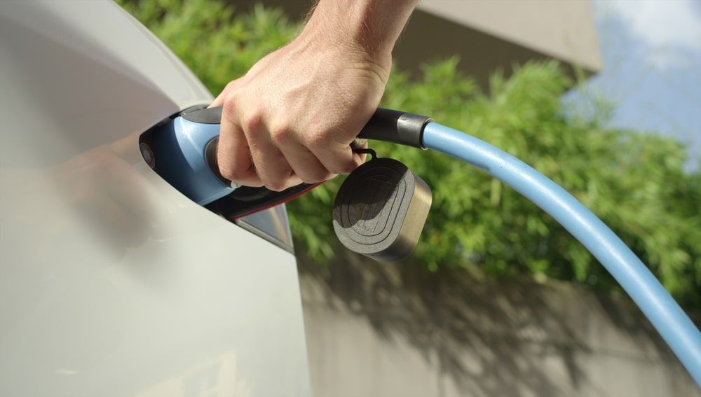 Recharge de voiture électrique : branchement du câble bleu Main branchant un câble de recharge bleu sur une voiture électrique blanche dans un cadre extérieur verdoyant.