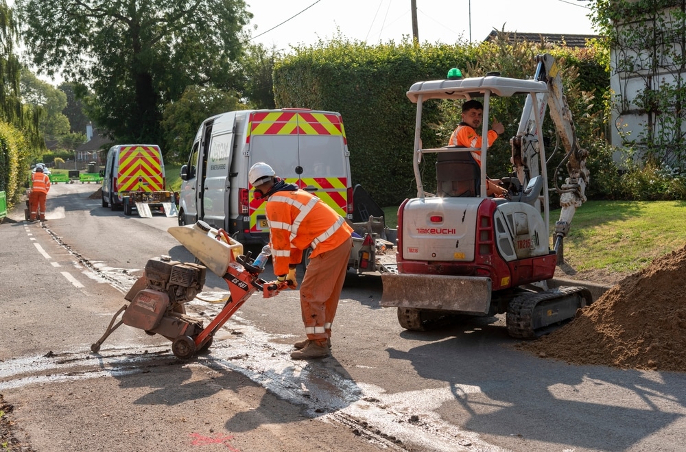 Des ouvriers en tenue haute visibilité utilisent une scie à sol et une mini-pelle Takeuchi pour des travaux routiers.