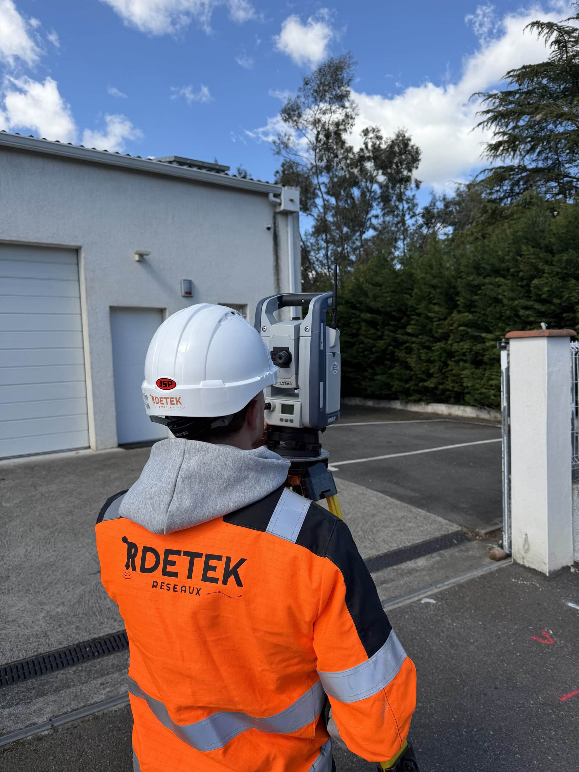 Technicien RDETEK Réseaux utilisant une station totale pour relevé topographique devant un bâtiment.