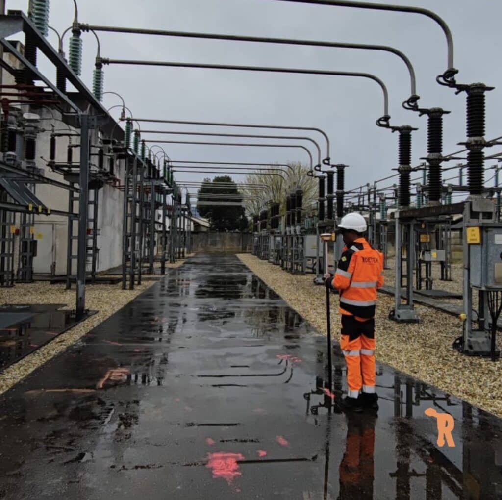 Relevés Topographiques en Poste Électrique Haute Tension Technicien effectuant des relevés topographiques de précision dans un poste électrique haute tension sous un ciel gris.