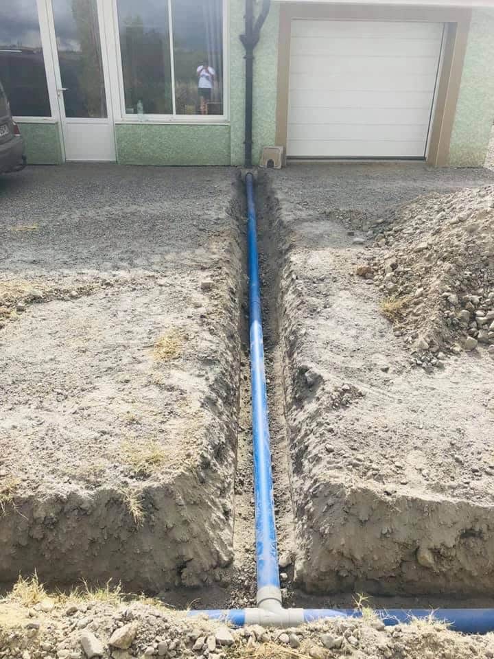 Installation drainage : Pose de tuyaux eaux de pluie Installation d'un tuyau de drainage bleu dans une tranchée de terre devant une maison avec garage et porte blanche.