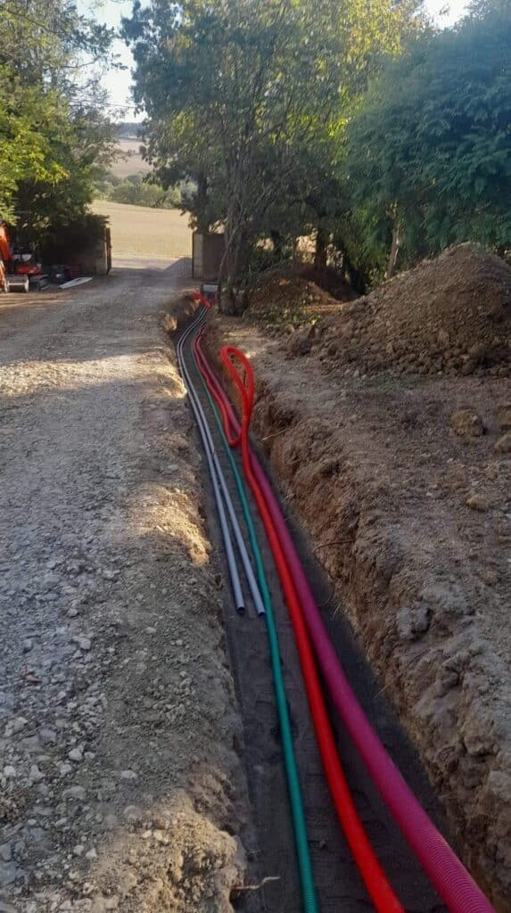 Installation de gaines pour réseaux enterrés en tranchée Tranchée de chantier avec gaines colorées (rouge, rose, vert) et tuyaux gris pour l'installation de réseaux enterrés.