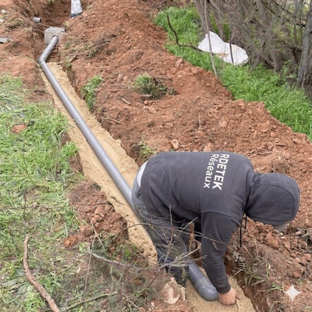 Un professionnel de RDETEK Réseaux installe un tuyau d'évacuation gris dans une tranchée ensablée en extérieur.